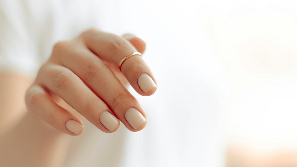 Nahaufnahme einer Hand mit hellbeige lackierten Nägeln und einem goldenen Ring auf dem Mittelfinger.